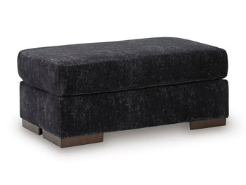 Ashley Furniture Whitmore Manor Ottoman in beige fabric, showing a plush, tufted top and wooden block legs from an angled view.