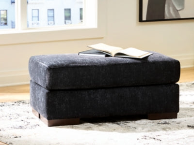 Ashley Furniture Whitmore Manor Ottoman with plush fabric and wooden legs, shown in a cozy living room setting.