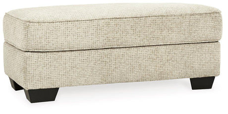 Ashley Furniture Monaghan Ottoman in neutral fabric with wooden legs, showcasing its plush cushioned top and stylish design.