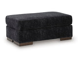 Ashley Furniture Whitmore Manor Ottoman in beige fabric, showing a plush, tufted top and wooden block legs from an angled view.