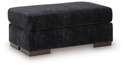 Ashley Furniture Whitmore Manor Ottoman in neutral fabric, shown from an angled side view with plush cushioning and wooden legs.